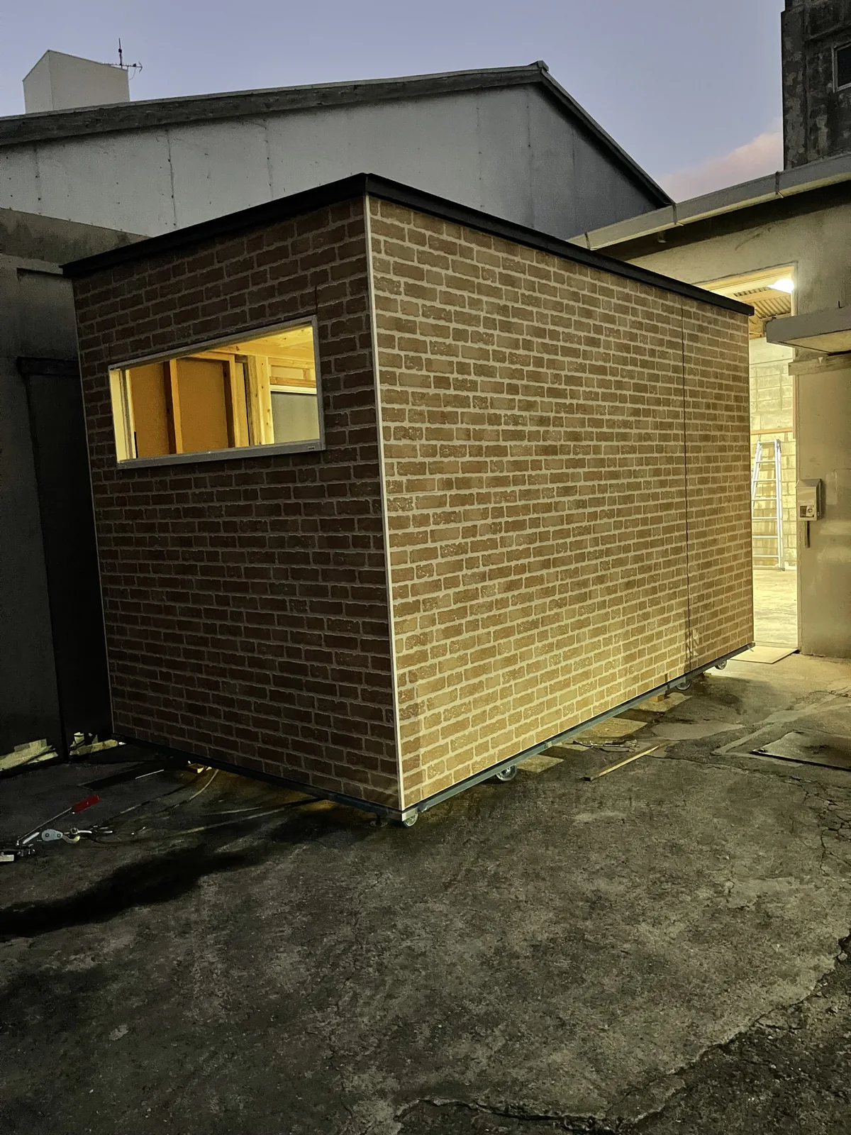 夕暮れの空の下、窓から暖かな明かりが漏れるレンガ調の外壁の小屋